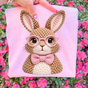 May include: A light pink sweatshirt featuring a crocheted bunny wearing pink glasses and a bow tie. The bunny has brown fur with white and pink accents. The design is centered on the sweatshirt.