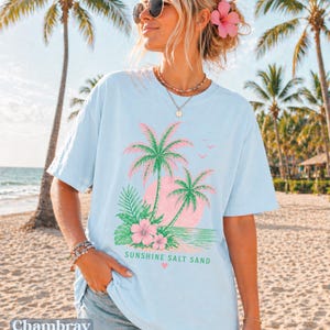 May include: Pale blue t-shirt with a tropical design of palm trees, flowers, and the words "Sunshine Salt Sand." The shirt has short sleeves and a relaxed fit, ideal for a beach holiday.