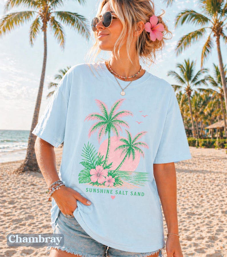 May include: Pale blue t-shirt with a tropical design of palm trees, flowers, and the words "Sunshine Salt Sand." The shirt has short sleeves and a relaxed fit, ideal for a beach holiday.