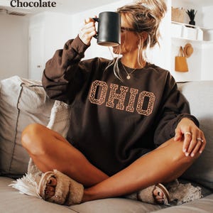 May include: A dark chocolate-coloured sweatshirt featuring the word "OHIO" in a leopard print design. A person is holding a black mug. The sweatshirt is part of a clothing collection.