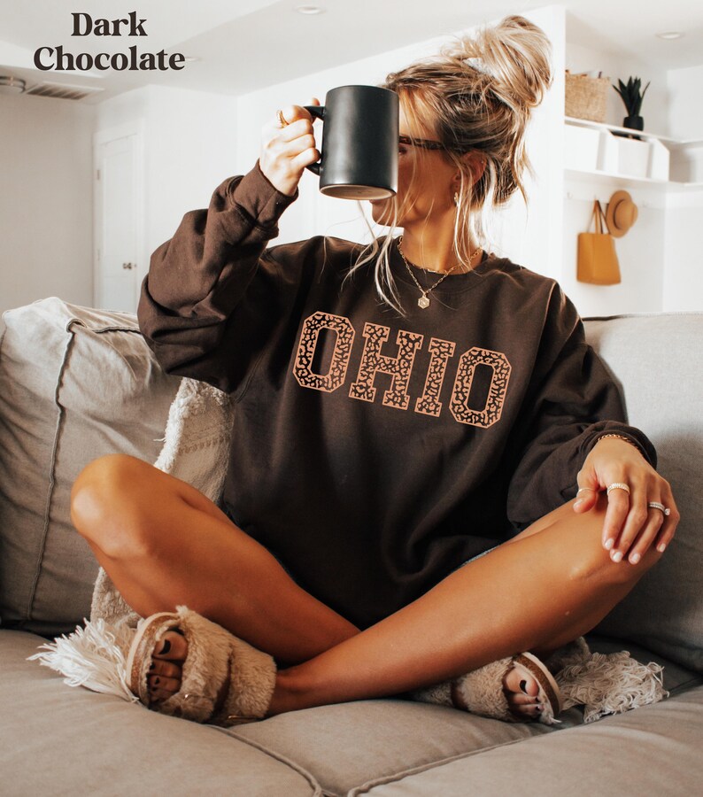 May include: A dark chocolate-coloured sweatshirt featuring the word "OHIO" in a leopard print design. A person is holding a black mug. The sweatshirt is part of a clothing collection.