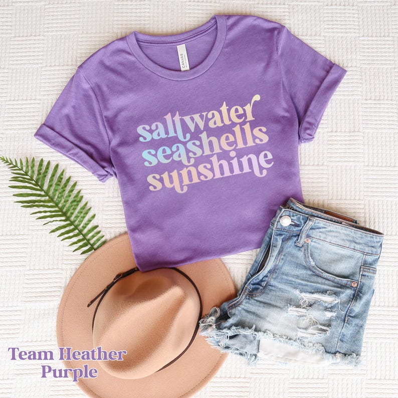 May include: A purple t-shirt with the words "saltwater seashells sunshine" in a pastel gradient. The shirt is paired with denim shorts, a tan hat, and a fern. The text "Team Heather Purple" is at the bottom.