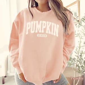 Puede incluir: Una sudadera de color melocot&oacute;n con las palabras "PUMPKIN SEASON" impresas en letras blancas. La sudadera es de estilo cuello redondo y tiene mangas largas. La palabra "Peachy" est&aacute; impresa en la esquina inferior derecha.