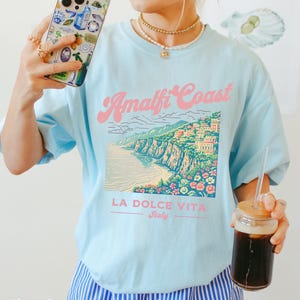 May include: A light blue t-shirt with a graphic of the Amalfi Coast, Italy. The graphic features a colorful illustration of the coastline with buildings and flowers. The text "Amalfi Coast" is in pink and the text "LA DOLCE VITA Italy" is in black.
