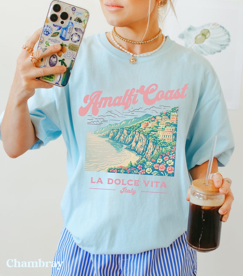 May include: A light blue t-shirt with a graphic of the Amalfi Coast, Italy. The graphic features a colorful illustration of the coastline with buildings and flowers. The text "Amalfi Coast" is in pink and the text "LA DOLCE VITA Italy" is in black.