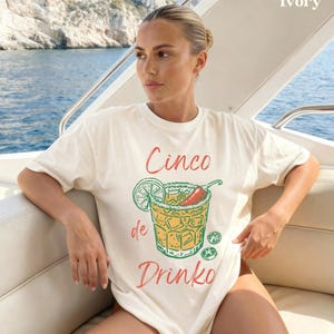 May include: Ivory-colored t-shirt with a Cinco de Drinko graphic. The graphic features a cocktail with a lime wedge, chili pepper, and salt rim. The text "Cinco de Drinko" is in red and green. The shirt is worn on a boat.