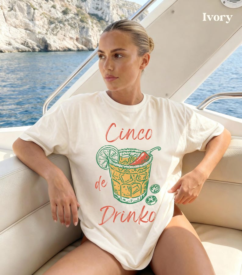 May include: Ivory-colored t-shirt with a Cinco de Drinko graphic. The graphic features a cocktail with a lime wedge, chili pepper, and salt rim. The text "Cinco de Drinko" is in red and green. The shirt is worn on a boat.