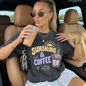 Puede incluir: Camiseta gris oscuro con el texto "Sunshine iced & Coffee all day every day" en amarillo y morado. La camiseta presenta un gr&aacute;fico de una taza de caf&eacute;. La persona lleva gafas de sol y joyas y sostiene un caf&eacute; helado.