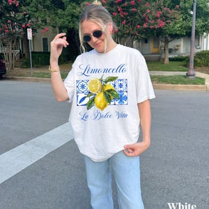 Pu&ograve; includere: Maglietta bianca con una grafica di limoni e piastrelle blu e bianche. Il testo "Limoncello La Dolce Vita" &egrave; stampato sulla maglietta.