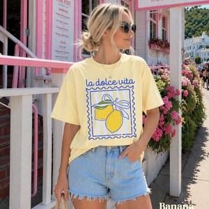 Pu&ograve; includere: T-shirt giallo chiaro con la scritta "la dolce vita" e un disegno di limoni. Abbinato a pantaloncini di jeans azzurri. Lo sfondo mostra un edificio rosa e fiori.