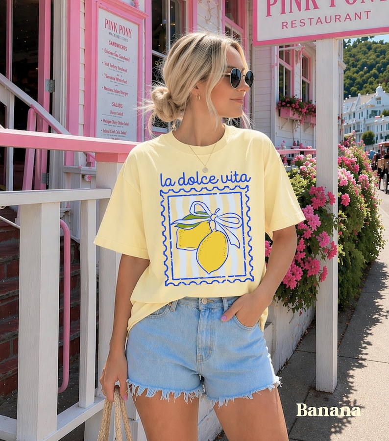 Pu&ograve; includere: T-shirt giallo chiaro con la scritta "la dolce vita" e un disegno di limoni. Abbinato a pantaloncini di jeans azzurri. Lo sfondo mostra un edificio rosa e fiori.