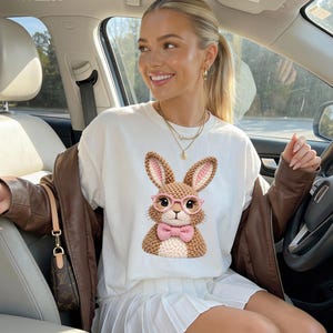 May include: White t-shirt featuring a cartoon bunny wearing pink glasses and a bow tie. The bunny has brown and pink crochet details. The word "White" is printed in the top left corner of the image.