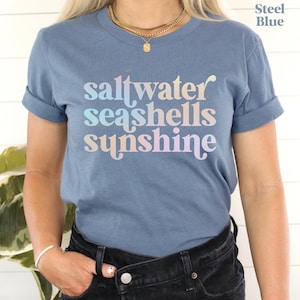 May include: Steel blue t-shirt with rolled-up sleeves. The shirt features the words "saltwater seashells sunshine" in a pastel rainbow font. A gold necklace and bracelet are visible.