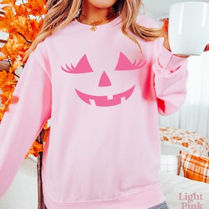 May include: A light pink long-sleeve sweatshirt featuring a dark pink graphic of a jack-o'-lantern face with eyelashes. The design includes two triangular eyes with lashes, a small triangular nose, and a wide, jagged, smiling mouth.