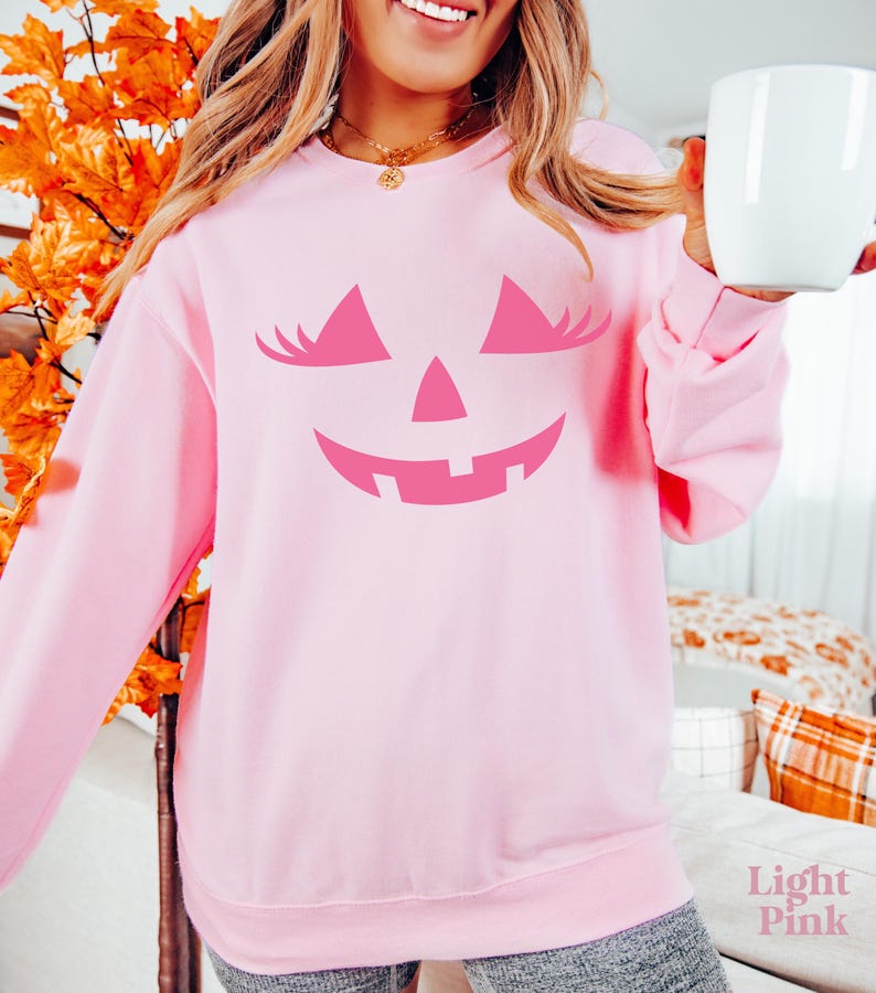 May include: A light pink long-sleeve sweatshirt featuring a dark pink graphic of a jack-o'-lantern face with eyelashes. The design includes two triangular eyes with lashes, a small triangular nose, and a wide, jagged, smiling mouth.