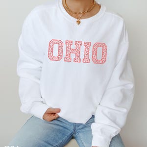 May include: White sweatshirt featuring the word "OHIO" in a coral leopard print. The long-sleeved top is made of a soft material and is suitable for casual wear. The word "White" is printed at the bottom of the image.