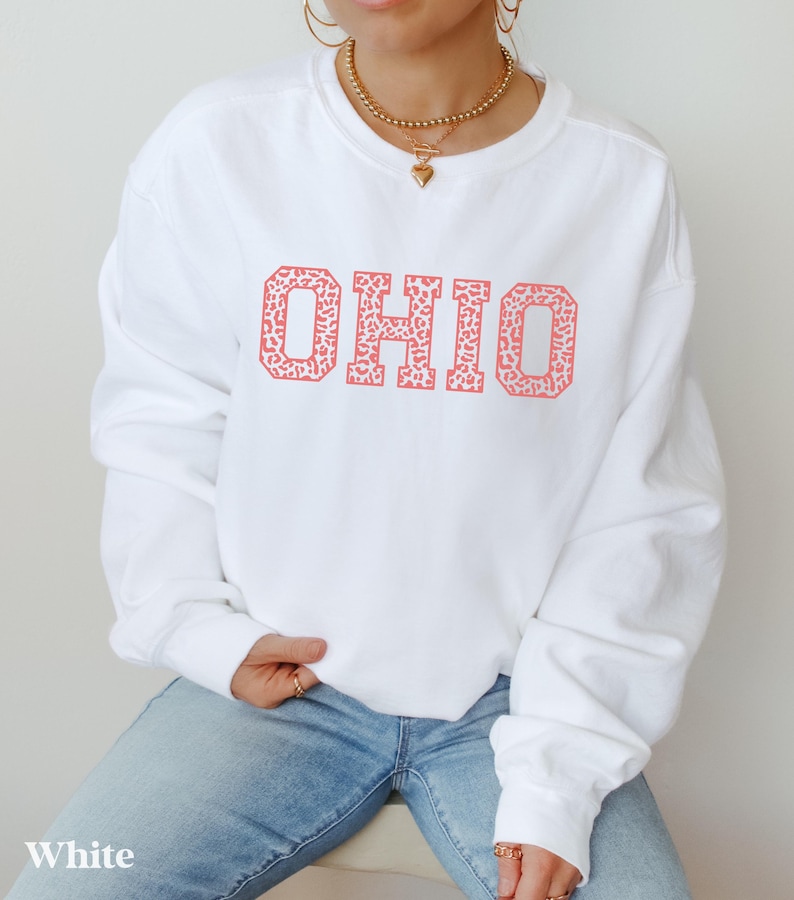 May include: White sweatshirt featuring the word "OHIO" in a coral leopard print. The long-sleeved top is made of a soft material and is suitable for casual wear. The word "White" is printed at the bottom of the image.