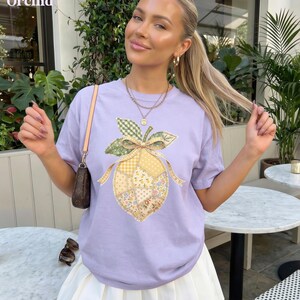 May include: A lilac t-shirt with a lemon patchwork design. The lemon has floral, gingham, and solid fabric patterns, with a green and yellow bow. The word "Orchid" is in the top left corner.