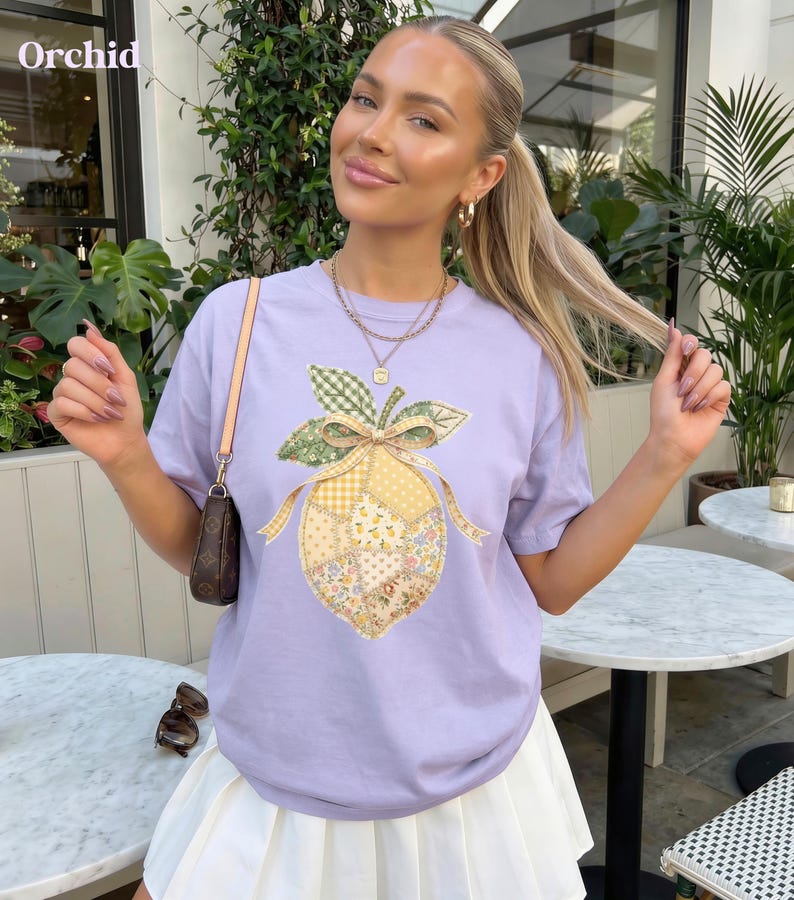 May include: A lilac t-shirt with a lemon patchwork design. The lemon has floral, gingham, and solid fabric patterns, with a green and yellow bow. The word "Orchid" is in the top left corner.
