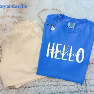 May include: A blue t-shirt with the words "HELLO sunshine" in white and gold, alongside beige shorts. A gold chain necklace with a pendant is also visible. The items are arranged on a soft, textured surface, creating a relaxed aesthetic.