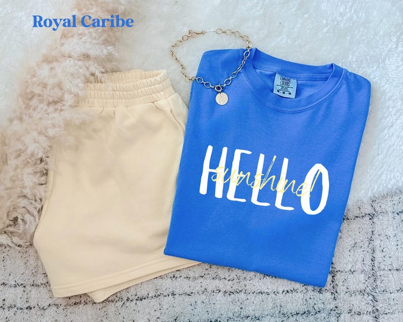 May include: A blue t-shirt with the words "HELLO sunshine" in white and gold, alongside beige shorts. A gold chain necklace with a pendant is also visible. The items are arranged on a soft, textured surface, creating a relaxed aesthetic.