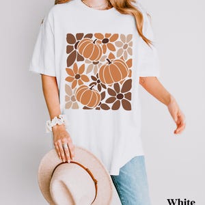 May include: White t-shirt featuring a fall-themed design with pumpkins and flowers in shades of brown and orange. The shirt has a relaxed fit and short sleeves. The word "White" is printed on the bottom right.