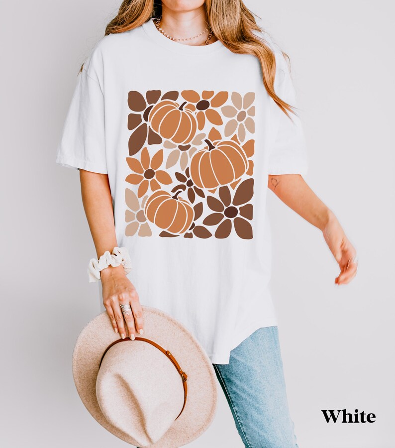 May include: White t-shirt featuring a fall-themed design with pumpkins and flowers in shades of brown and orange. The shirt has a relaxed fit and short sleeves. The word "White" is printed on the bottom right.