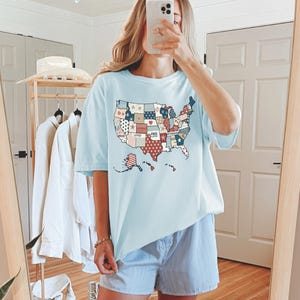 May include: A light blue t-shirt with a patchwork design of the United States map. The map is made up of different colored squares with words like "Brave", "Loved", and "Free" written on them. The shirt is being worn by a person with a light blue and white striped pair of shorts.