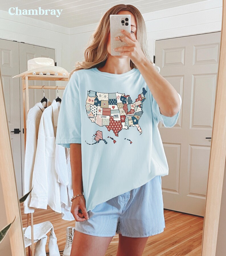 May include: A light blue t-shirt with a patchwork design of the United States map. The map is made up of different colored squares with words like "Brave", "Loved", and "Free" written on them. The shirt is being worn by a person with a light blue and white striped pair of shorts.