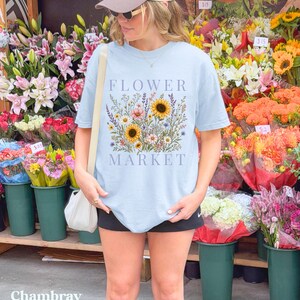May include: A light blue t-shirt featuring a graphic print of a colorful wildflower bouquet with sunflowers and the text "FLOWER MARKET" in a serif font. The shirt has short sleeves and a crew neckline, worn with a beige baseball cap and a white bag.