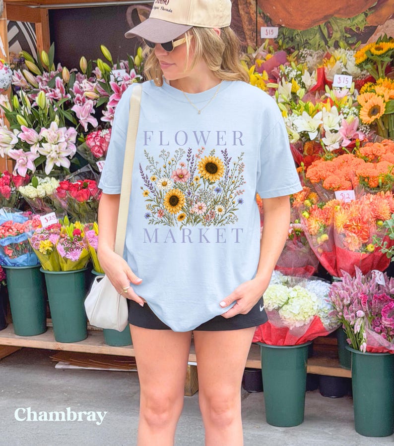 May include: A light blue t-shirt featuring a graphic print of a colorful wildflower bouquet with sunflowers and the text "FLOWER MARKET" in a serif font. The shirt has short sleeves and a crew neckline, worn with a beige baseball cap and a white bag.