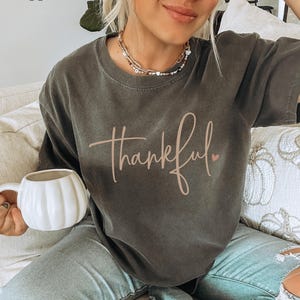 May include: Pepper-colored t-shirt with the word "thankful" in a cursive font and a small pink heart. The shirt is a dark gray color. A white pumpkin-shaped mug is held in the hand.