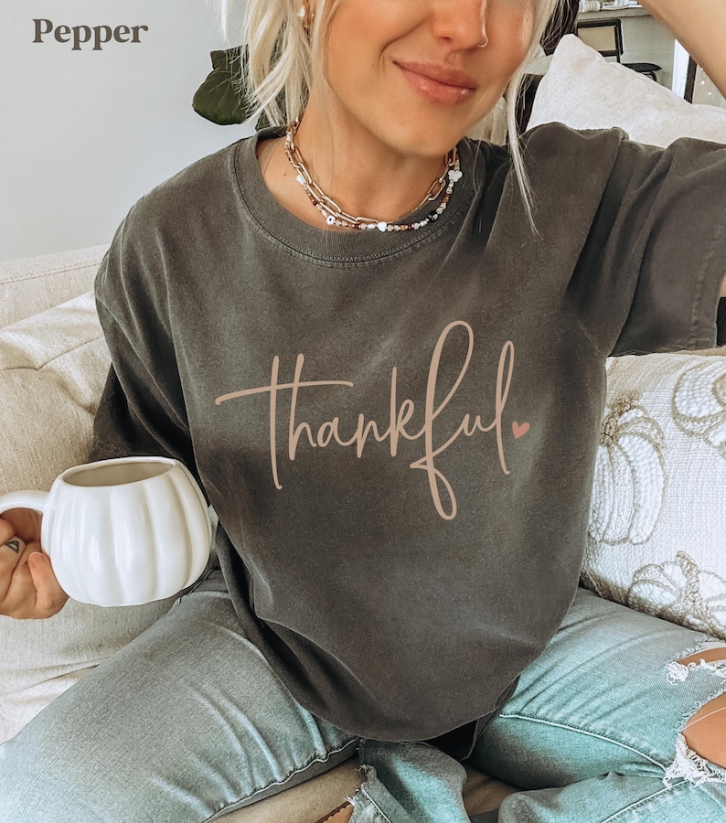 May include: Pepper-colored t-shirt with the word "thankful" in a cursive font and a small pink heart. The shirt is a dark gray color. A white pumpkin-shaped mug is held in the hand.