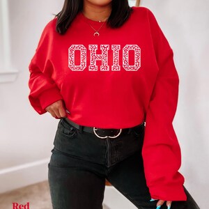 May include: A red sweatshirt displaying the word "OHIO" in white, with a patterned design within the letters. The sweatshirt is worn with black jeans and a black belt with a silver buckle. The word "Red" is in the bottom left corner.