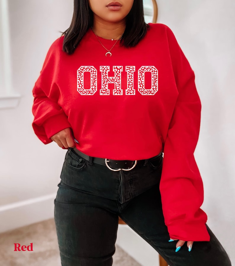 May include: A red sweatshirt displaying the word "OHIO" in white, with a patterned design within the letters. The sweatshirt is worn with black jeans and a black belt with a silver buckle. The word "Red" is in the bottom left corner.