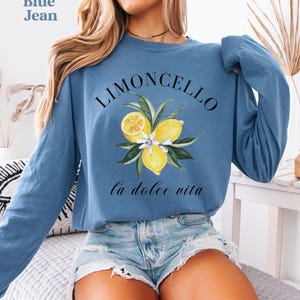 May include: Blue jean long-sleeve sweatshirt with a watercolor design. The design features the word "Limoncello" above a cluster of lemons and leaves, with the phrase "la dolce vita" below. The sweatshirt is a light blue color.