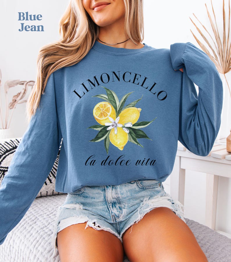 May include: Blue jean long-sleeve sweatshirt with a watercolor design. The design features the word "Limoncello" above a cluster of lemons and leaves, with the phrase "la dolce vita" below. The sweatshirt is a light blue color.