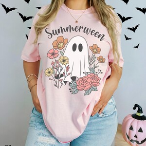 May include: A light pink t-shirt with the word "Summerween" in black script above a white ghost illustration surrounded by colorful flowers. The shirt is worn with blue jeans. A pink pumpkin with a jack-o'-lantern face is on the right.