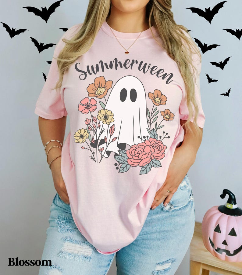 May include: A light pink t-shirt with the word "Summerween" in black script above a white ghost illustration surrounded by colorful flowers. The shirt is worn with blue jeans. A pink pumpkin with a jack-o'-lantern face is on the right.