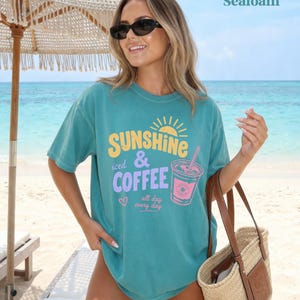 Puede incluir: Una camiseta turquesa con las palabras "Sunshine iced & Coffee" en amarillo y morado, junto con un gr&aacute;fico de una bebida de caf&eacute;. La camiseta se lleva en una playa con un bolso de paja.