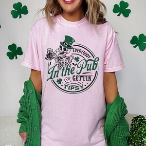 skeleton pub crawl shirt for your St. Patrick's Day party that has a skeleton holding a beer wearing a green Leprechaun hat with shamrocks saying Everybody in the Pub Gettin' Tipsy on a Comfort Colors shirt in the color blossom pink