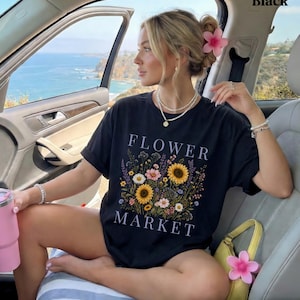 May include: A black short-sleeved t-shirt featuring a graphic print of a colorful wildflower bouquet with sunflowers and daisies. The text "FLOWER MARKET" is printed in white capital letters above and below the floral arrangement.