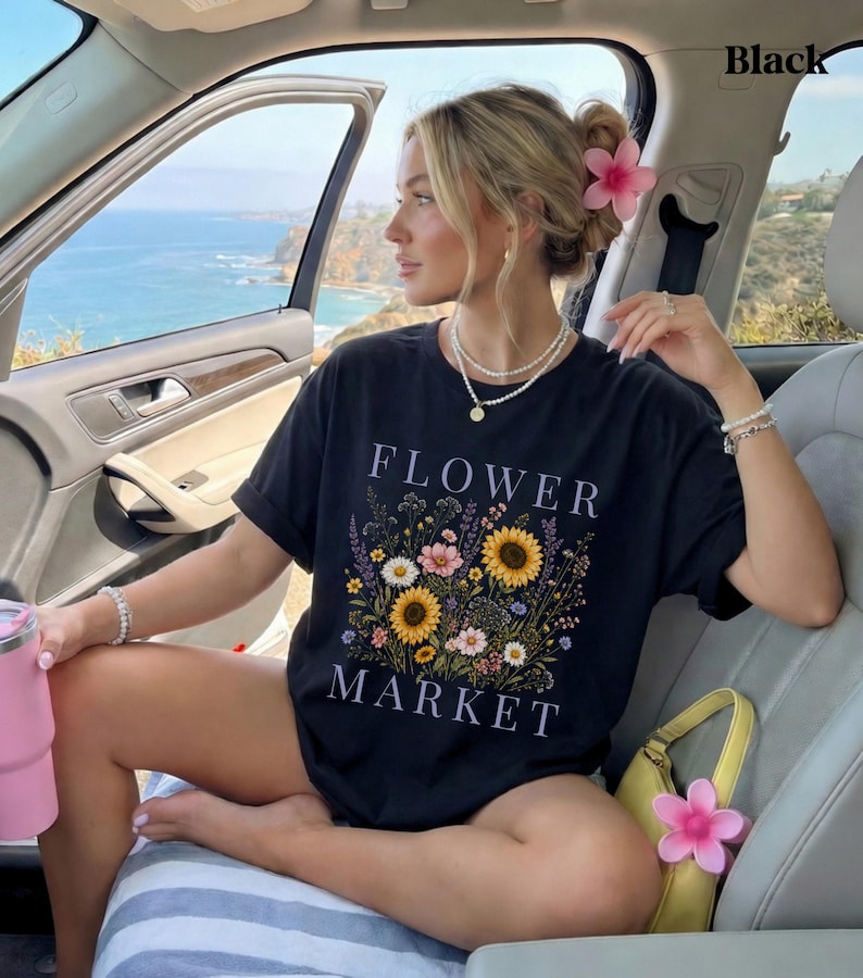 May include: A black short-sleeved t-shirt featuring a graphic print of a colorful wildflower bouquet with sunflowers and daisies. The text "FLOWER MARKET" is printed in white capital letters above and below the floral arrangement.