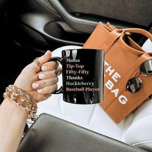 May include: Black ceramic mug with the words "Mama Tip-Top Fifty-Fifty Thanks Huckleberry Baseball Player" in white and pink lettering. A brown tote bag and sunglasses are in the background.