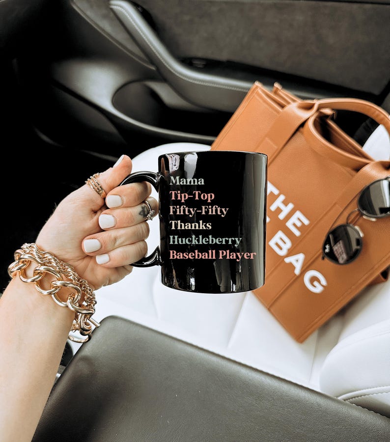 May include: Black ceramic mug with the words "Mama Tip-Top Fifty-Fifty Thanks Huckleberry Baseball Player" in white and pink lettering. A brown tote bag and sunglasses are in the background.