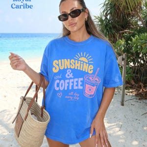 Puede incluir: Una camiseta azul con el texto "SUNSHINE iced & COFFEE all day every day" y un gr&aacute;fico de una bebida de caf&eacute;. La camiseta se usa en una playa con un bolso de mano de paja.