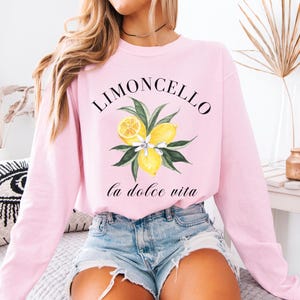 May include: A light pink sweatshirt with the word "Limoncello" above a watercolor illustration of lemons and leaves. Below the illustration, the words "La Dolce Vita" are written in a script font. The word "Blossom" is in the top left corner.