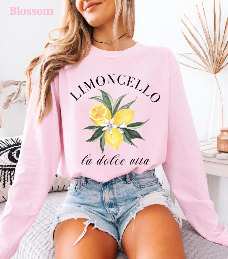 May include: A light pink sweatshirt with the word "Limoncello" above a watercolor illustration of lemons and leaves. Below the illustration, the words "La Dolce Vita" are written in a script font. The word "Blossom" is in the top left corner.