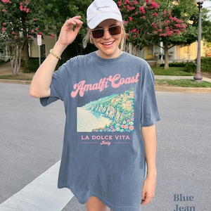 May include: A blue oversized t-shirt with a graphic print of the Amalfi Coast, Italy. The text "Amalfi Coast" is in pink and the text "La Dolce Vita Italy" is in black.