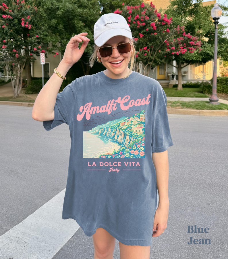 May include: A blue oversized t-shirt with a graphic print of the Amalfi Coast, Italy. The text "Amalfi Coast" is in pink and the text "La Dolce Vita Italy" is in black.